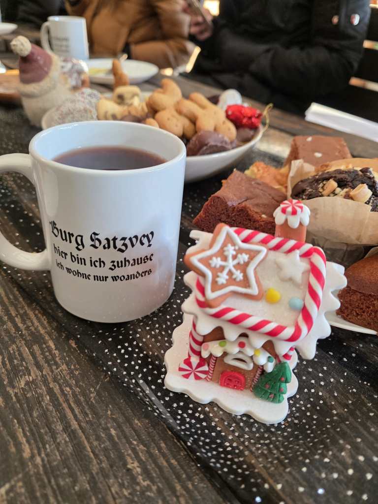 Tasse Glühwein mit der Aufschrift "Burg Satzvey, hier bin ich zuhause, ich wohne nur woanders", ein mini Lebkuchenhaus und viele weitere süße Leckereien